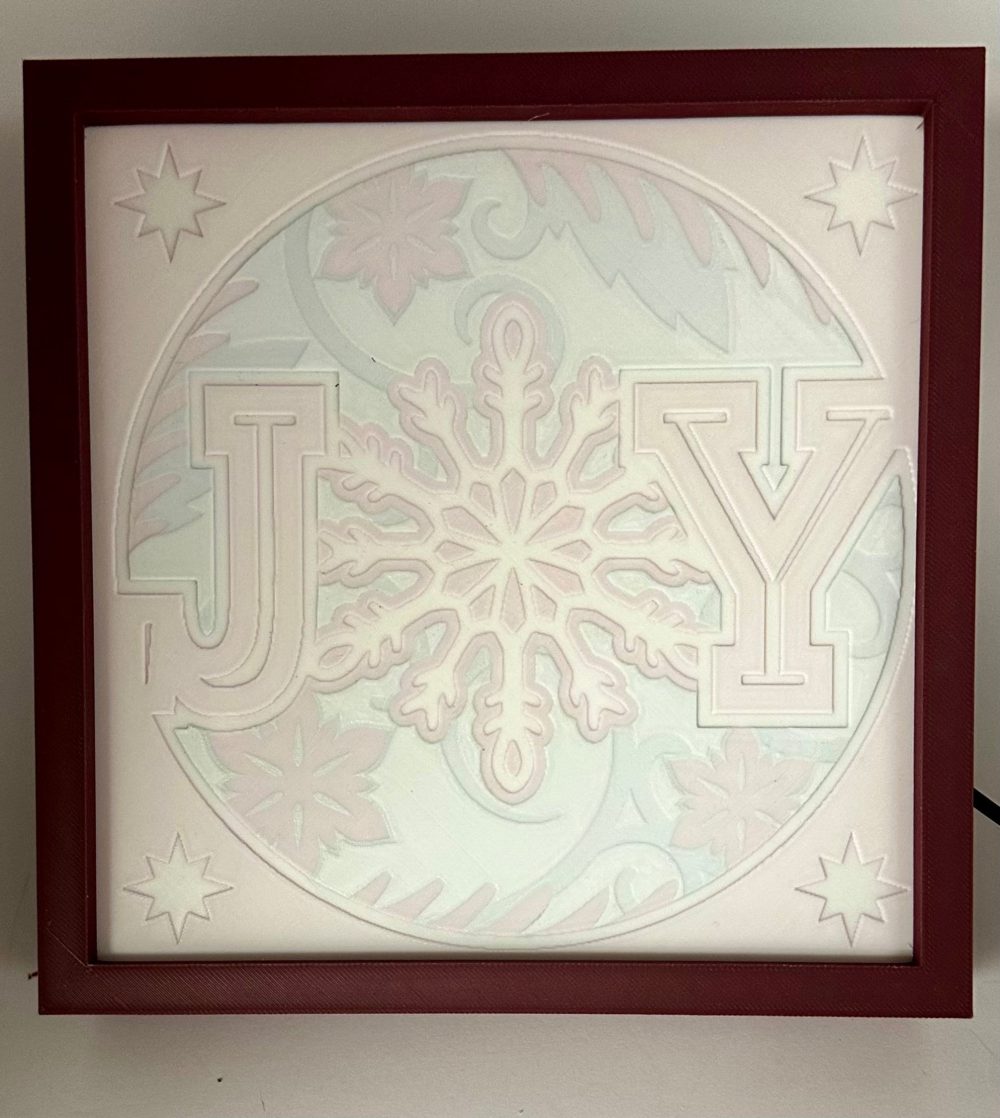 JOY Christmas light decoration featuring a festive snowflake design with embossed letters on a square panel.